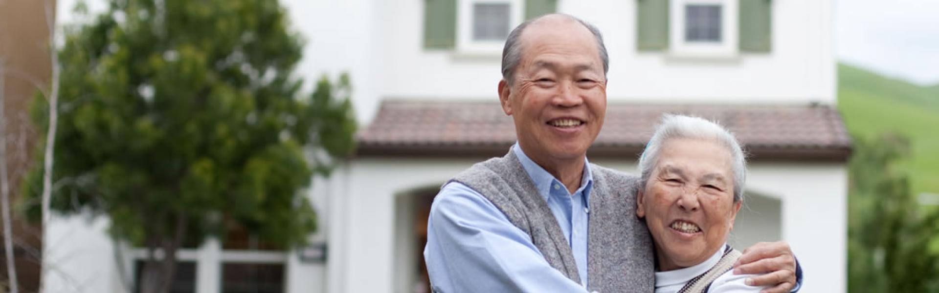 Older Asian couple embracing and posing for a picture in front of their home